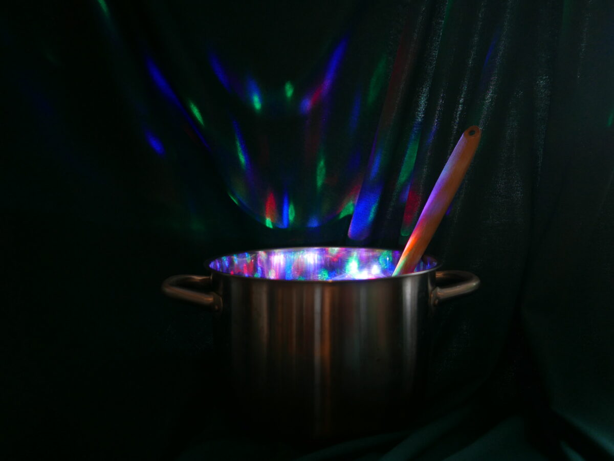 Das Bild zeigt einen Kochtopf mit Holzlöffel vor dunkelgrünem Vorhang. Aus dem Topf kommen bunte Partylichter, die auch auf dem Vorhang zu erkennen sind