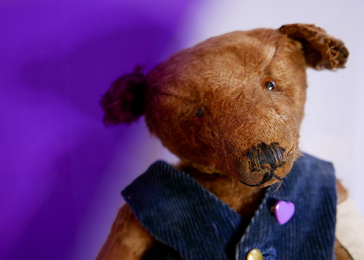 Kopf und Oberkörper eines alten Teddys mit Gebrauchsspuren. Er hat kurzes hellbraunes Fell und trägt eine selbstgenähte dunkelblaue Weste mit Messing-Knöpfen und einer Herz-Brosche. Er guckt traurig.